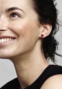 Orecchini a forma di cuore rossi con accenti trasparenti, posizionati contro l'orecchio di un modello. Gli orecchini sono realizzati in un materiale lucido, che esalta il colore vibrante.