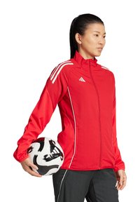 Rote Sportjacke mit weißen Akzenten und Logo, mit hohem Kragen und Reißverschluss, gehalten mit einem schwarz-weißen Fußball.