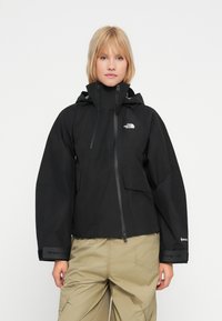 Ženska nosi črno vodoodporno jakno North Face s kapuco in bež tovorne hlače, stoji ob navadni beli ozadju.