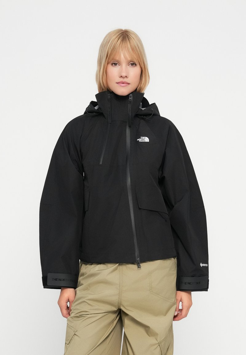 Ženska nosi črno vodoodporno jakno North Face s kapuco in bež tovorne hlače, stoji ob navadni beli ozadju.