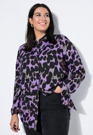 Nainen hymyilee, hänellä on päällään violetti ja musta abstrakti kuvioinen nappipaita ja mustat teksturoidut housut, seisoo valkoista taustaa vasten.