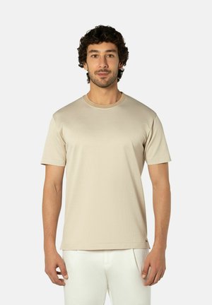 Mann mit kurzen lockigen Haaren, der ein beiges kurzärmliges T-Shirt und weiße Hose trägt und vor einem einfarbigen hellen Hintergrund steht.