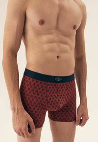 Red boxer shorts with a geometric pattern, made from soft fabric. Features a contrast navy waistband with branded tag at the back.