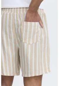 Shorts rayés beiges en tissu léger avec une taille élastique et une poche sur le côté, agrémentés de rayures verticales crème et marron clair.