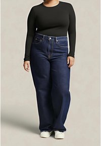 Jean denim bleu taille haute avec un design classique à cinq poches, associé à un haut noir à manches longues et des chaussures blanches.