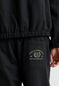 Îmbrăcăminte sport neagră, cu un material texturat, tiv elastic și o patch logo proeminentă pe buzunarul lateral.