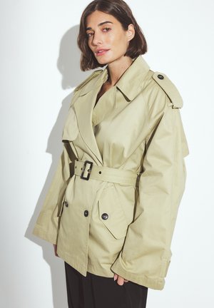 Licht kaki trenchcoat met een ceintuur in de taille, grote klepzakken en schouderepauletten. Beschikt over een klassieke kraag en knoopsluiting.