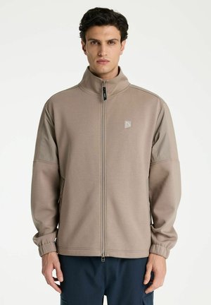 Mann mit kurzen dunklen Haaren, der eine beige Jacke mit Reißverschluss und elastischen Bündchen sowie dunkle Hose trägt, steht vor einem weißen Hintergrund.