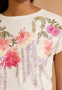 Cream-colored top featuring floral embroidery in pink, white, and purple tones, with detailed leaves and hanging vine patterns.