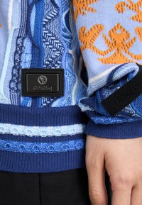 Blå striktrøje med geometriske mønstre i nuancer af blå og orange, ribbede manchetter og et sort logo-patch på siden.