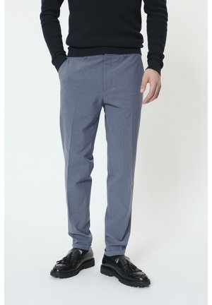 Man draagt slanke lichtblauwe broek, zwarte trui met lange mouwen, zwarte sokken en zwarte leren loafers met franjes, staand met handen deels in de zakken.