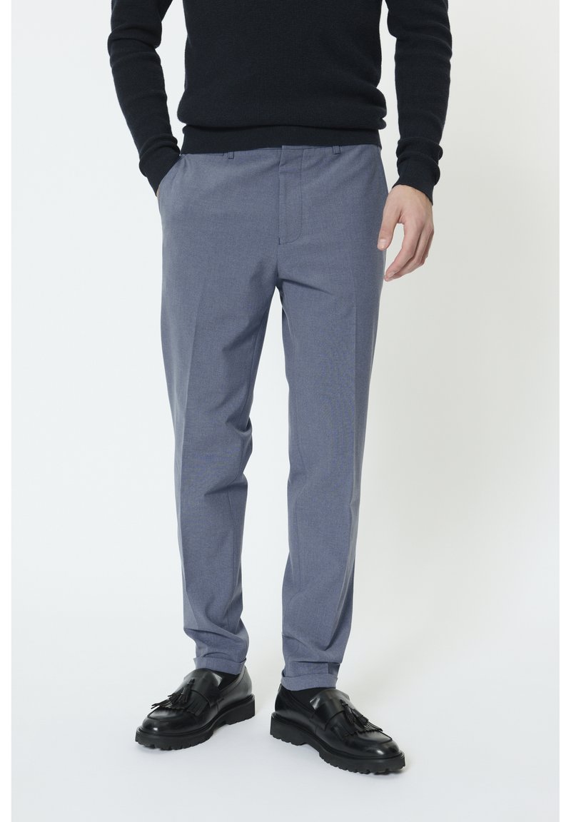 Hombre con pantalones ajustados azul claro, suéter negro de manga larga, calcetines negros y mocasines de cuero negros con borlas, de pie con las manos parcialmente en los bolsillos.