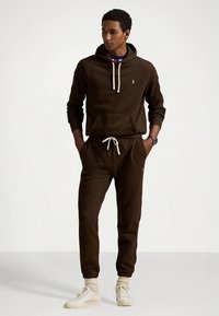 Brun bomullshettegenser med kremfargede snorer, matchende joggebukser og en liten logo. Modellen har på lyse sneakers og ribbestrømper.