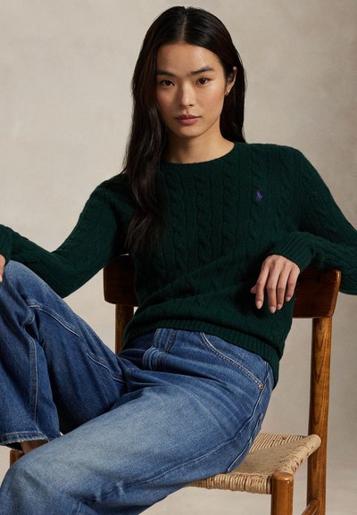 Giovane donna con capelli lunghi e scuri che indossa un maglione a trecce verde scuro e jeans blu, seduta rilassata su una sedia di legno.