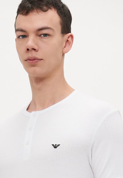 Baltas Henley tipo marškinėliai su tekstūruota medžiaga, turintys tris mygtukus priekyje ir mažą juodą logotipą kairiajame krūtinės pusėje.