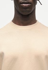 Άτομο με σκούρο δέρμα που φοράει ανοιχτό μπεζ μπλουζάκι με στρογγυλή λαιμόκοψη, εμφανιζόμενο από το λαιμό μέχρι το πάνω μέρος του στήθους, σε λευκό απλό φόντο.