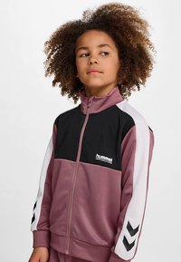 Veste de performance Hummel en rose clair avec panneaux en mesh noir, accents blancs et fermeture éclair sur le devant. Comprend un motif chevron sur les manches.