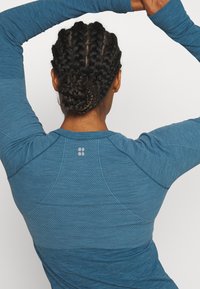 Top azul de manga larga con tejido texturizado, que presenta un diseño ajustado, detalles en los hombros sin costuras en contraste y un logo sutil cerca del cuello.