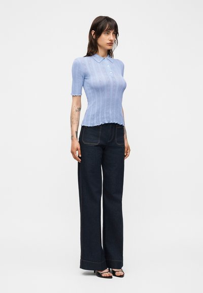 Γυναίκα με ανοιχτό μπλε πλεκτό πόλο με κοντά μανίκια, σκούρο φαρδύ τζιν και μαύρα σανδάλια ανοιχτών δαχτύλων, στέκεται μπροστά σε λευκό φόντο.