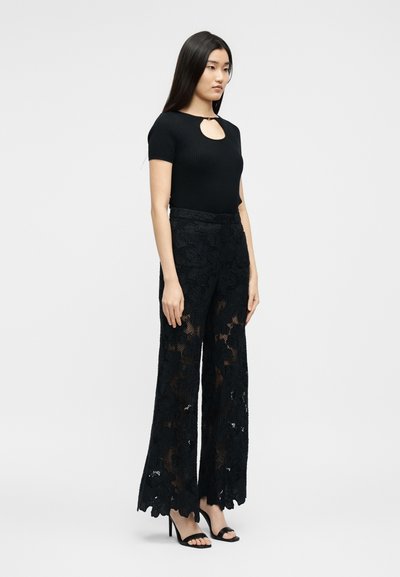 Γυναίκα όρθια, φοράει μαύρο τοπ με κοντά μανίκια και άνοιγμα στο λαιμό, καθώς και μαύρο παντελόνι φαρδιά δαντέλα με ψηλοτάκουνα σανδάλια.