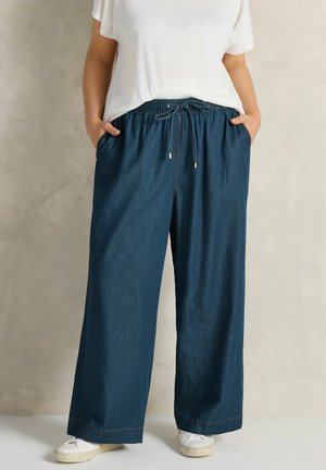 Osoba ubrana w luźne, granatowe dżinsowe spodnie z troczkiem w pasie, białą koszulkę, białe trampki, stojąca na tle jednolitego beżowego tła.