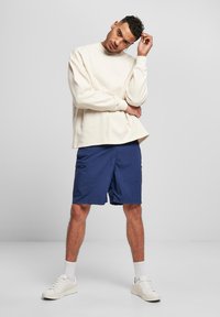 Hanorac crem cu mâneci lungi, asortat cu pantaloni scurți bleumarin cu un buzunar lateral. Adidași albi completează ținuta. Fundal neutru.