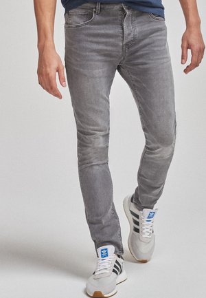 Persoon draagt strakzittende grijze spijkerbroeken en witte Adidas-sneakers met zwarte strepen, loopt naar voren tegen een effen achtergrond.