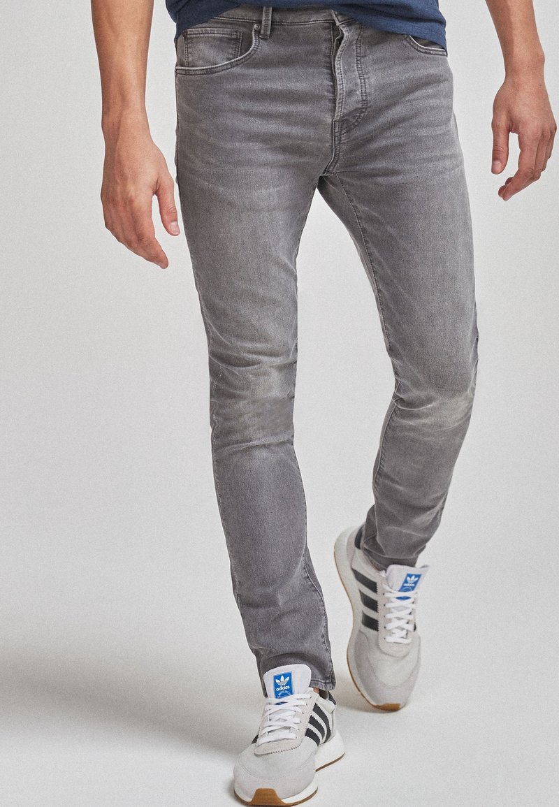 Personne portant un jean gris slim et des baskets Adidas blanches à rayures noires, marchant vers l'avant sur un fond uni.