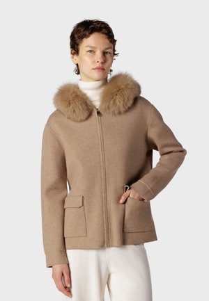 Kvinde iført en beige sweatcoat med fluffy pelskrave og hvide bukser, stående mod en ensfarvet lys grå baggrund.