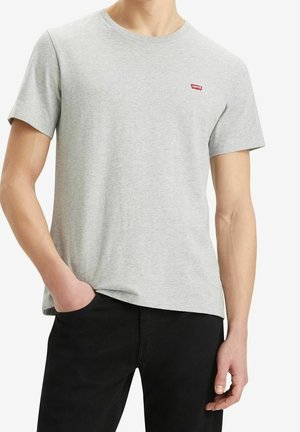 Mężczyzna w gładkim jasnoszarym krótkim rękawem t-shircie z małym czerwonym logo Levi's na piersi i czarnych spodniach, ręka w kieszeni.