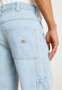 Pantalons cargo en denim bleu clair avec poches latérales et arrière. Présente un logo Dickies rouge, jaune et bleu sur la poche arrière. Design décontracté.