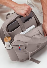 Grå ryggsäck med flera fack, en vattenflaskhållare och en tygetikett; har vadderade axelremmar och dragkedjeslutning.