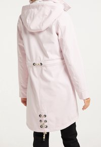 Manteau long imperméable rose clair avec capuche, taille cintrée et détail de laçage décoratif au dos. Tissu lisse et texturé.