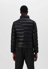 Zwarte pufferjack met een quilted ontwerp, hoge kraag en een gladde, glanzende textuur. Heeft een getailleerde pasvorm en elastische manchetten.