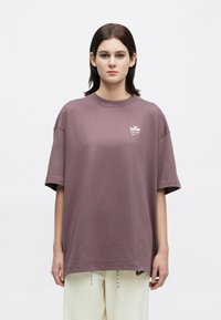 Mladostnik z ravnimi temnimi lasmi, oblečen v prevelik mauve majico z majhnim belim logotipom in smetano-bež hlačami, stoji pred enostavno ozadje.