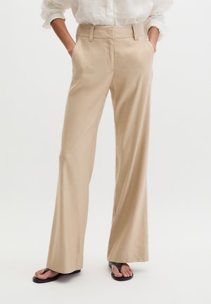 Beige wijde broek gedragen met een wit overhemd, handen in de zakken, gecombineerd met zwarte teensandaaltjes op een witte achtergrond.