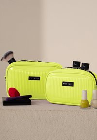 Zwei glänzende neon-gelbe Kosmetiktaschen mit gerippter Textur, schwarzen Akzenten und Reißverschlüssen. Der Inhalt umfasst Pinsel und Nagellackfläschchen.