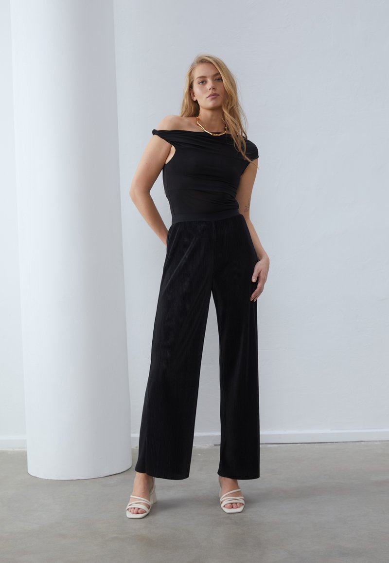 Top nero scollato abbinato a pantaloni ampi a coste neri. Sandali con cinturini chiari, semplice collana dorata, sfondo neutro.