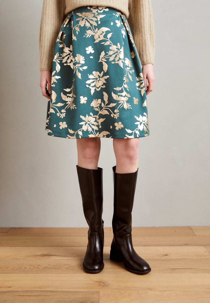 Femme portant une jupe bleu sarcelle à motifs floraux beige, un pull beige et des bottes en cuir marron foncé montant jusqu'aux genoux, debout sur un sol en bois.