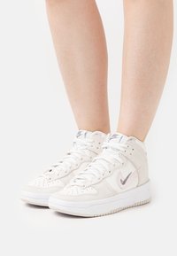 Vaaleansävyiset korkeavartiset tennarit, jotka on valmistettu nahasta ja mokasta. Ominaisuuksiin kuuluvat valkoiset nauhat, teksturoitu pohja ja hopeinen Nike-logo.