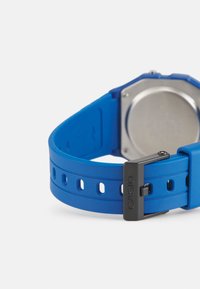 Bracelet de montre en silicone bleu avec plusieurs trous d'ajustement ; doté d'une boucle en métal noir et d'un boîtier circulaire argenté à l'extrémité.