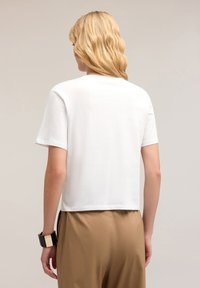 Camiseta corta de manga corta blanca en suave tejido de algodón, con un dobladillo recto y detalles de costura sutiles. Combinada con pantalones beis.