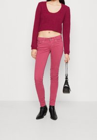 Suéter de ganchillo en rosa profundo, diseño recortado con un escote amplio, combinado con jeans ajustados rosas. Botines negros y un bolso pequeño completan el look.
