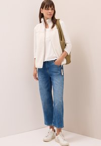 Kvinna som står i blå jeans, vita sneakers, vit långärmad tröja, krämfärgad fleeceväst och olivgrön axelväska mot en ljus vägg.