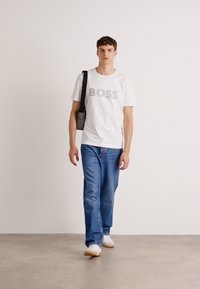 Witte t-shirt met "BOSS" in grijze letters, blauwe denim jeans en witte sneakers, accessorized met een zwarte schoudertas.