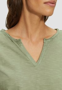 Grünes V-Ausschnitt-T-Shirt mit lockerer Passform und ausgefransten Kanten am Ausschnitt, aus weichem, leichtem Stoff mit subtiler Textur gefertigt.