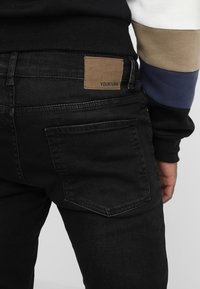 Svarta denimjeans med en brun läderlapp märkt "YOURTURN" på baksidan. Har enkla bakfickor och en figursydd design.
