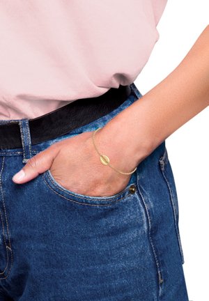 Gold chain bracelet with an oval charm, delicate design, shown on a hand resting in a denim pocket. Soft pink shirt and black belt visible.