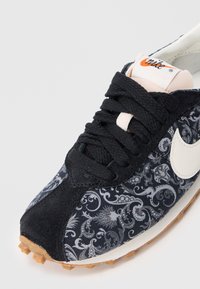 Sapatilhas pretas com parte superior em tecido apresentando um padrão cinza ornamentado, logo da Nike branco, atacadores pretos e uma sola de borracha castanha com tração.