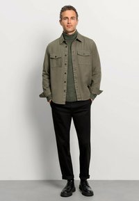 Olivgrünes Button-Up-Hemd mit zwei Brusttaschen, kombiniert mit einem dunkelgrünen Rollkragenpullover, schwarzen Hosen und schwarzen Schuhen.